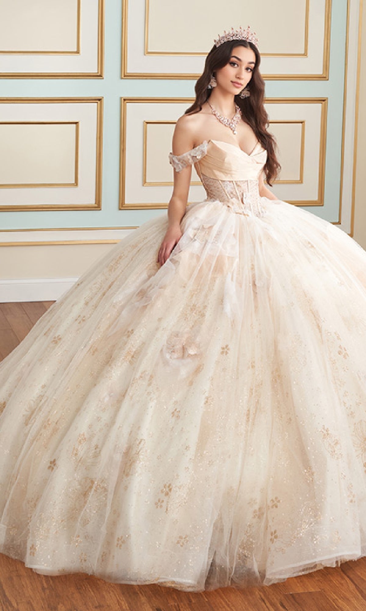A woman wearing the Quinceanera Dress PR30159 By Princesa, featuring a tiara, corset bodice, off-the-shoulder neckline, and full skirt embellished with 3D flowers poses indoors against a decorative wall.